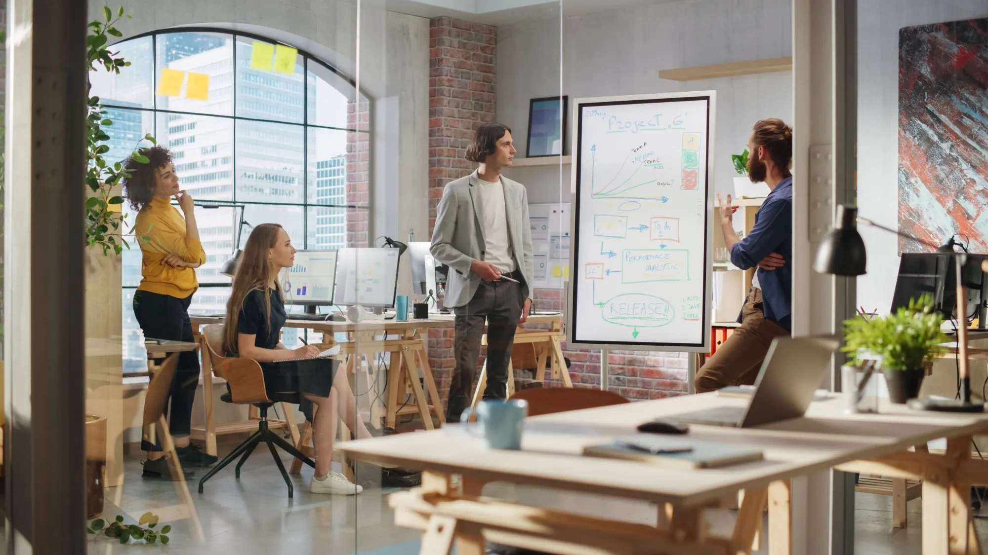 A team in a bright loft office gathered around an interactive display standing in portrait on its mobile stand: one of them is explaining a project flow drawn on it, with a growth chart, linked boxes and a release goal, while the others listen and take notes.